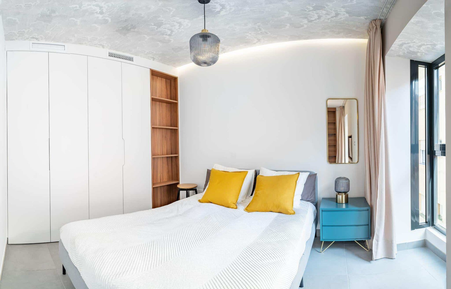 Clean bright bedroom with bed, duvet,side table, lamps,curtain, metal mirror, led indirect light and painted wallpaper on the ceiling.