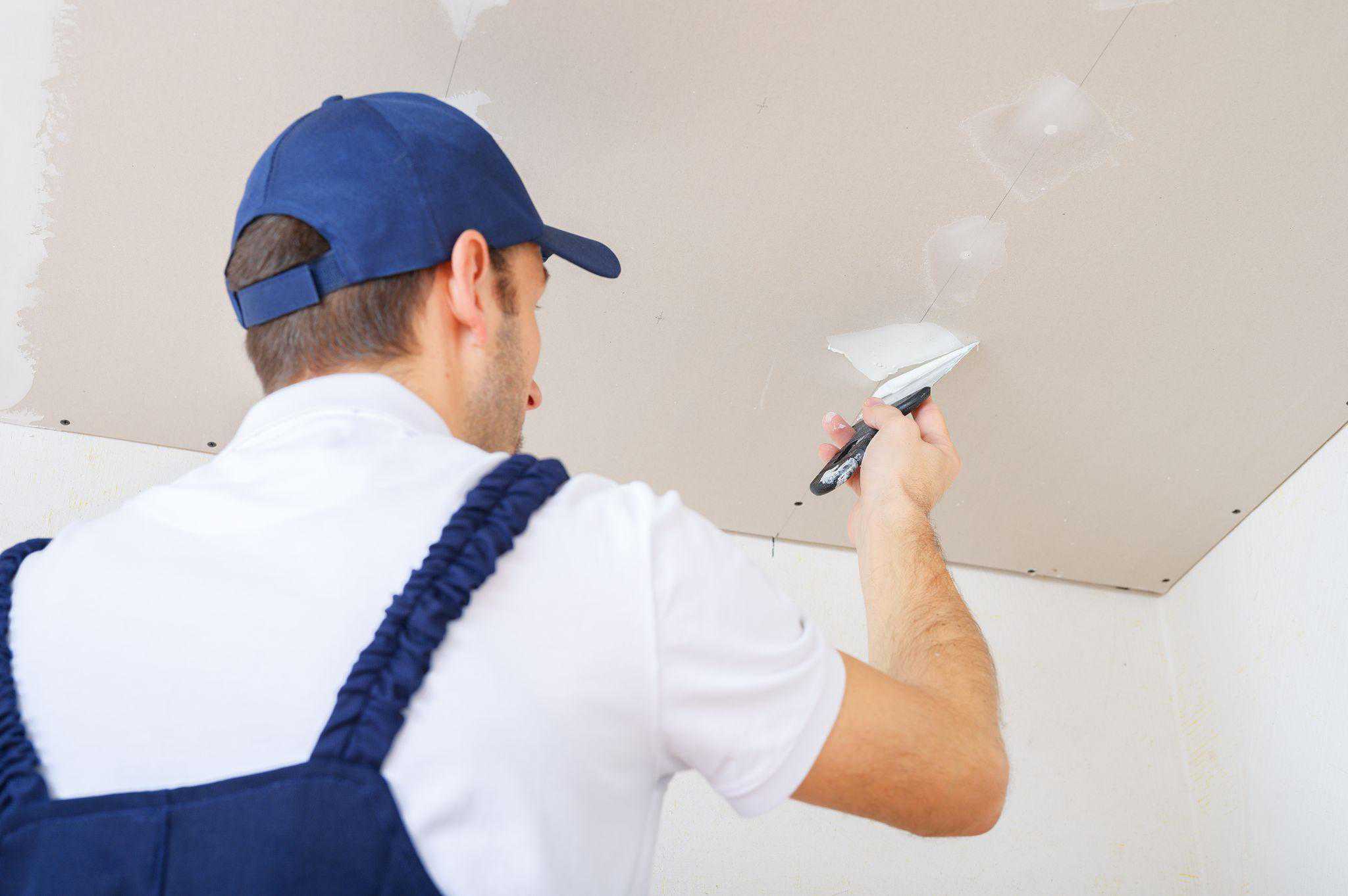 A uniformed worker puts putty on the caps of the screws on the ceiling made of drywall sheets.